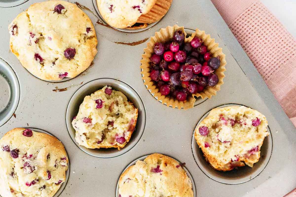 A muffin tin holds several baked berry muffins, one partially eaten muffin, and one cup filled with fresh berries. A pink towel is visible beside the tin.