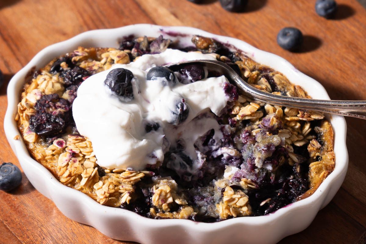 A baked oatmeal dish with blueberries and oats, topped with a dollop of yogurt, sits on a wooden surface with a spoon and scattered blueberries.
