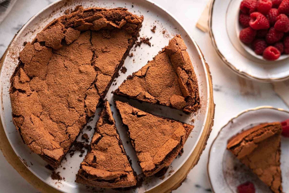 A round chocolate cake with a cracked top, partially sliced, sits on a plate with a bowl of raspberries and a plate with a cake slice nearby.
