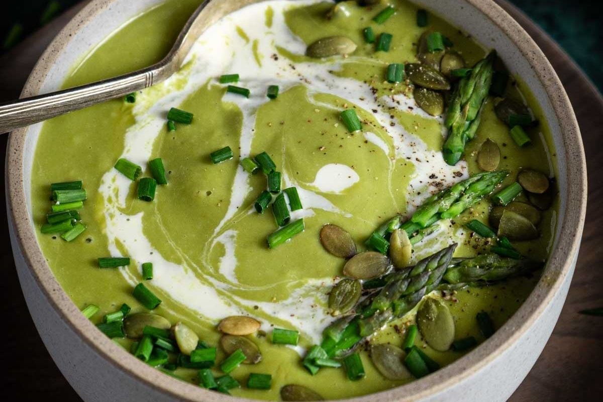 A bowl of green soup garnished with a swirl of cream, chopped chives, pumpkin seeds, black pepper, and asparagus tips, with a spoon resting inside.
