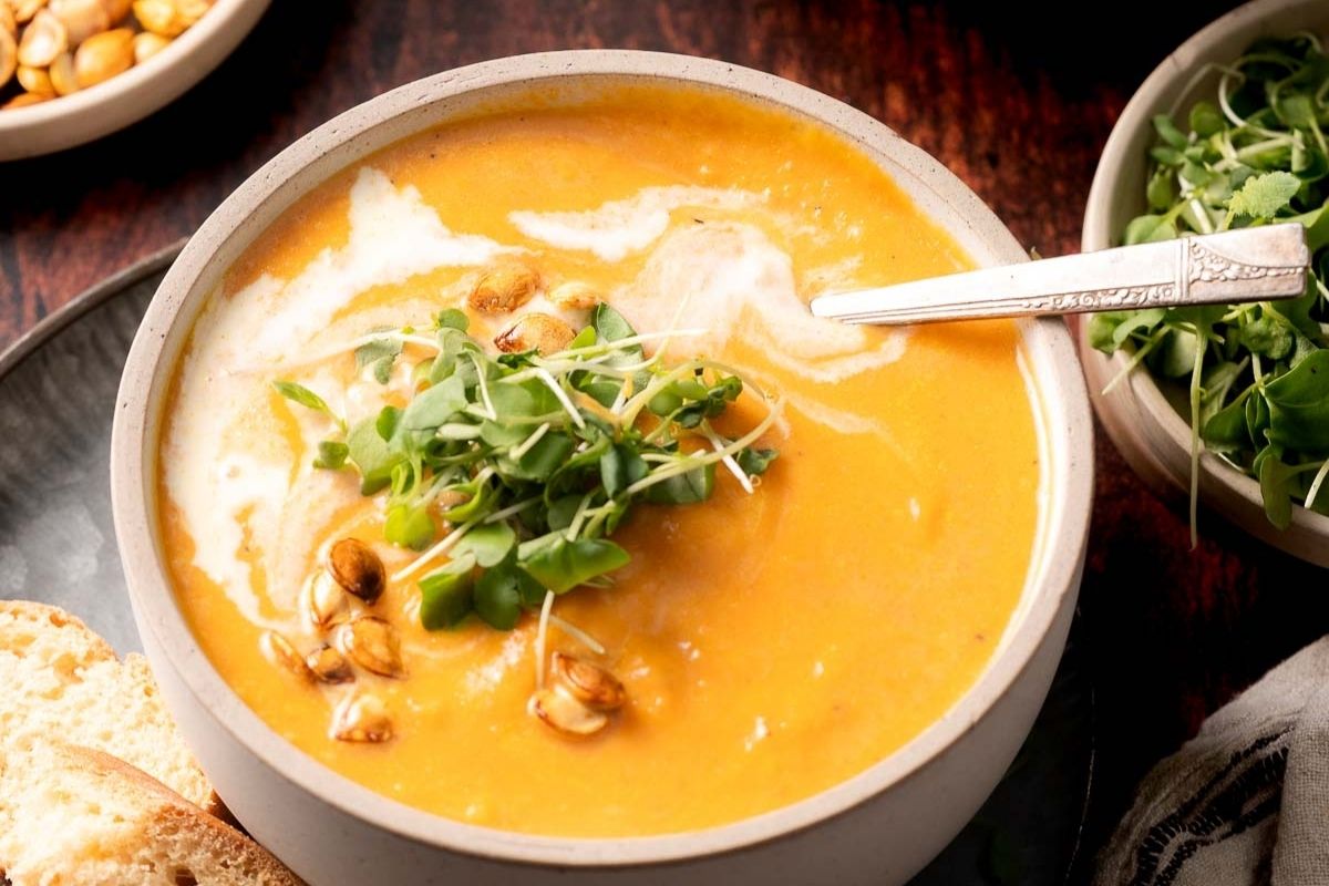 A bowl of creamy orange soup garnished with microgreens, roasted seeds, and a swirl of cream, with a spoon and bread on the side.