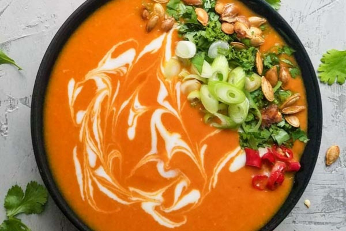 A bowl of orange soup with a swirl of cream, topped with sliced green onions, cilantro, pumpkin seeds, and chopped red chili.
