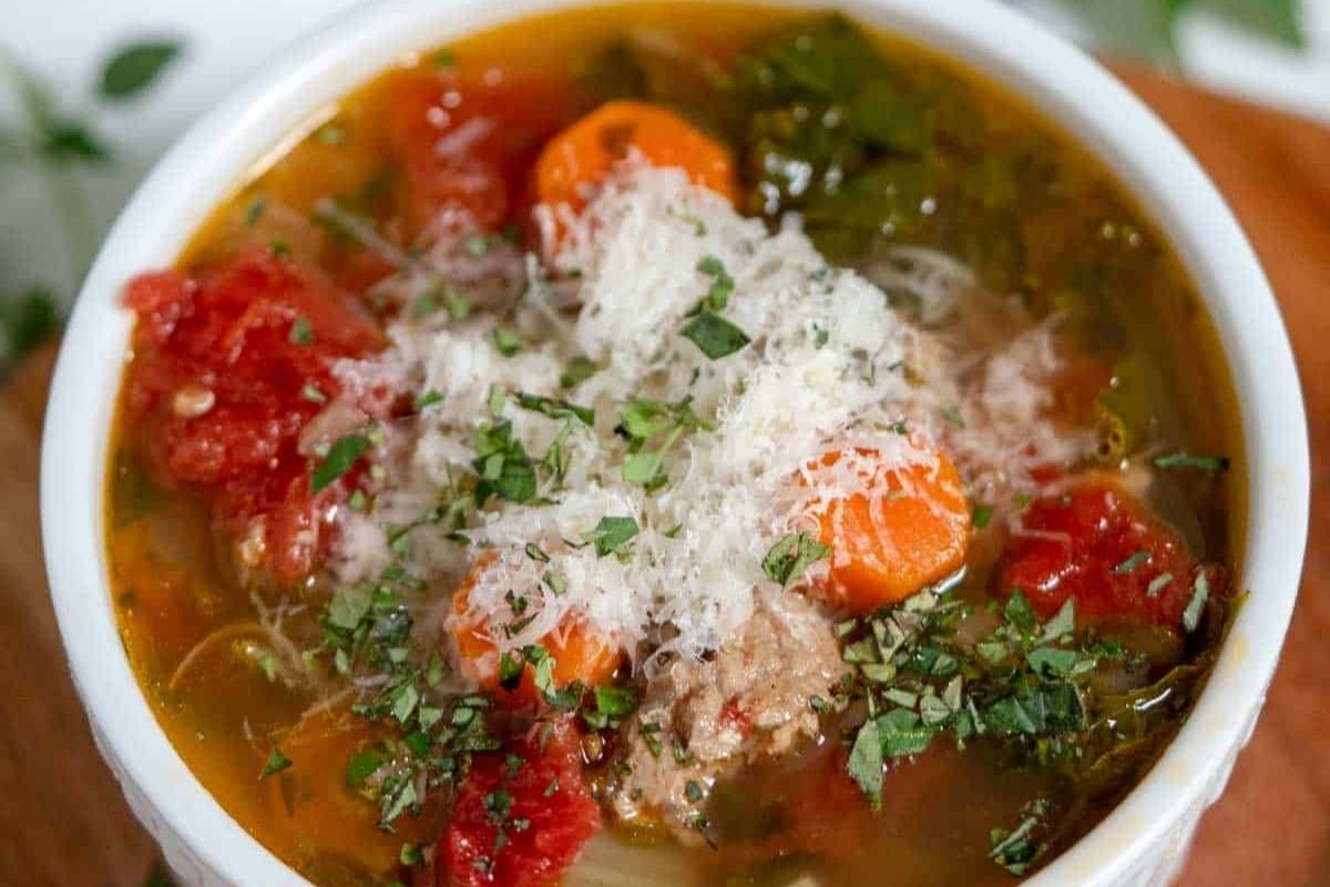 A bowl of soup with visible carrots, tomatoes, ground meat, leafy greens, grated cheese, and chopped herbs on top.