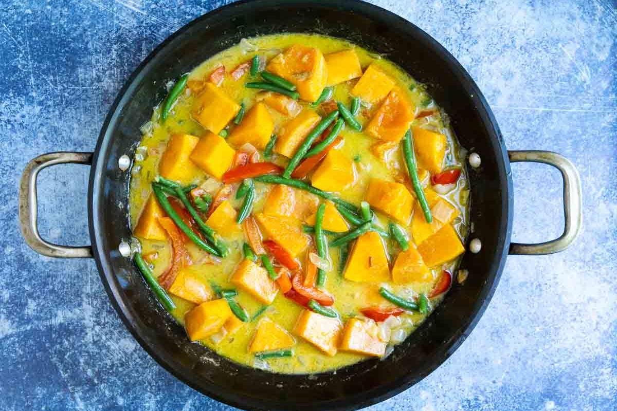 A pan filled with a yellow curry containing chunks of squash, green beans, and red bell peppers, placed on a blue textured surface.