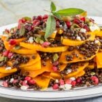 A plated salad of sliced butternut squash, black lentils, pomegranate seeds, pumpkin seeds, feta cheese, and fresh sage leaves as garnish.