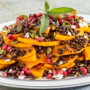A plated salad of sliced butternut squash, black lentils, pomegranate seeds, pumpkin seeds, feta cheese, and fresh sage leaves as garnish.