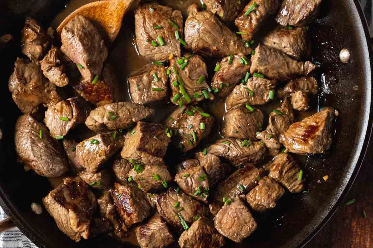 Cubed pieces of beef seared in a skillet, garnished with chopped chives, and stirred with a wooden spoon.