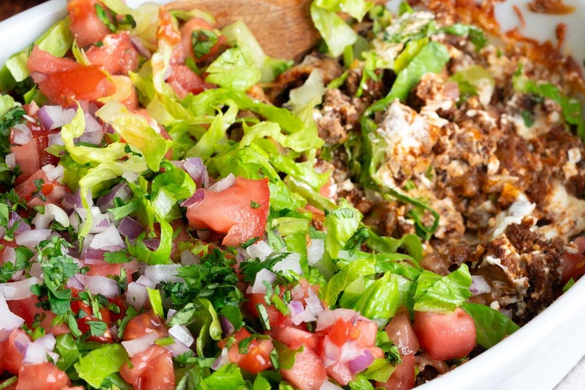 A close-up of a layered taco dip with ground beef, shredded lettuce, chopped tomatoes, red onions, and cilantro in a white dish.