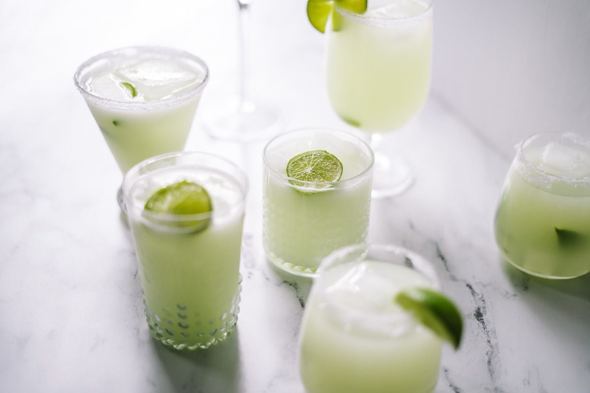 Five glasses filled with a light green beverage, each garnished with a slice of lime, are arranged on a white marble surface.
