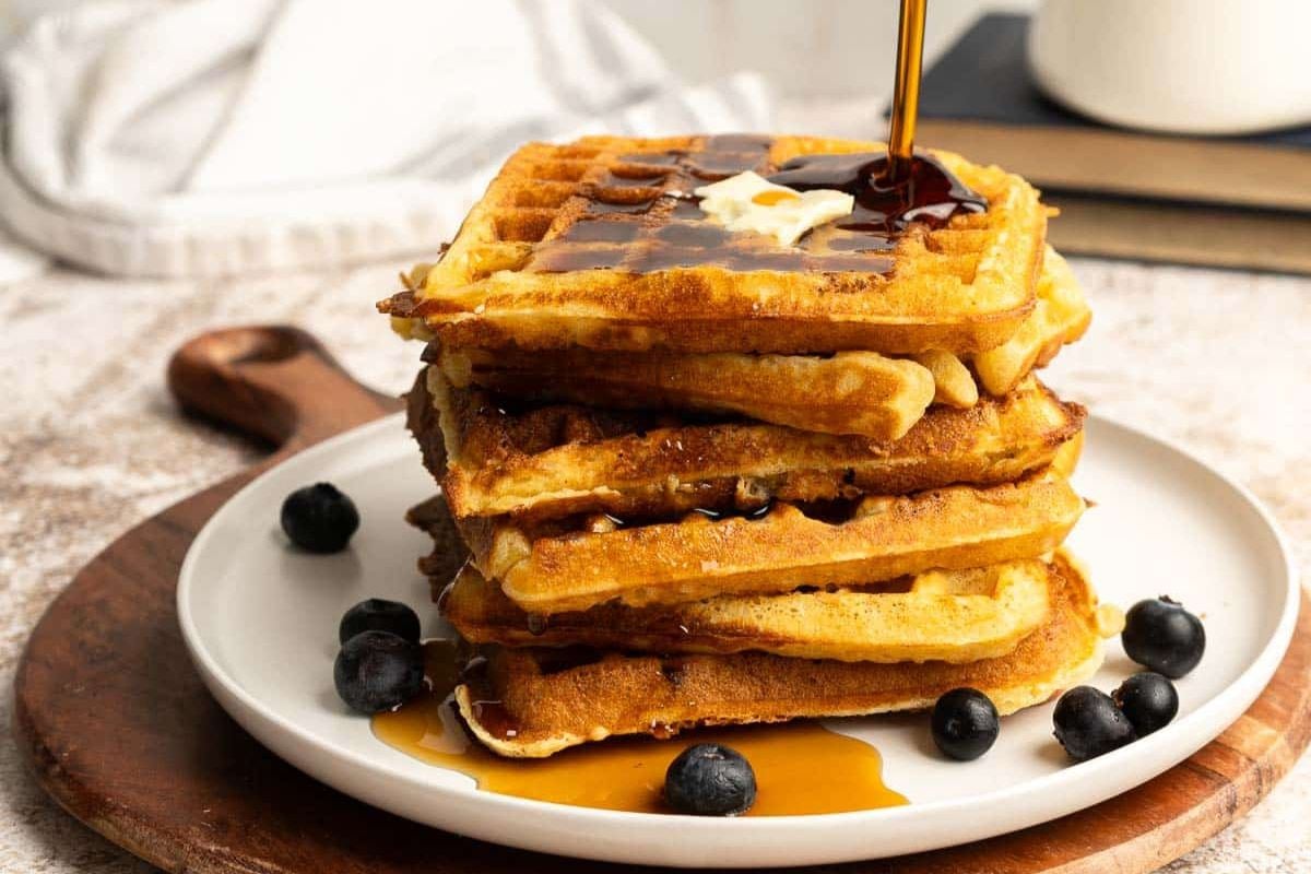 A stack of square waffles topped with butter and syrup on a white plate, surrounded by blueberries.