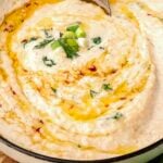 A bowl of creamy mashed potatoes garnished with chopped green onions, herbs, red pepper flakes, and drizzled with olive oil.