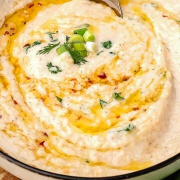 A bowl of creamy mashed potatoes garnished with chopped green onions, herbs, red pepper flakes, and drizzled with olive oil.