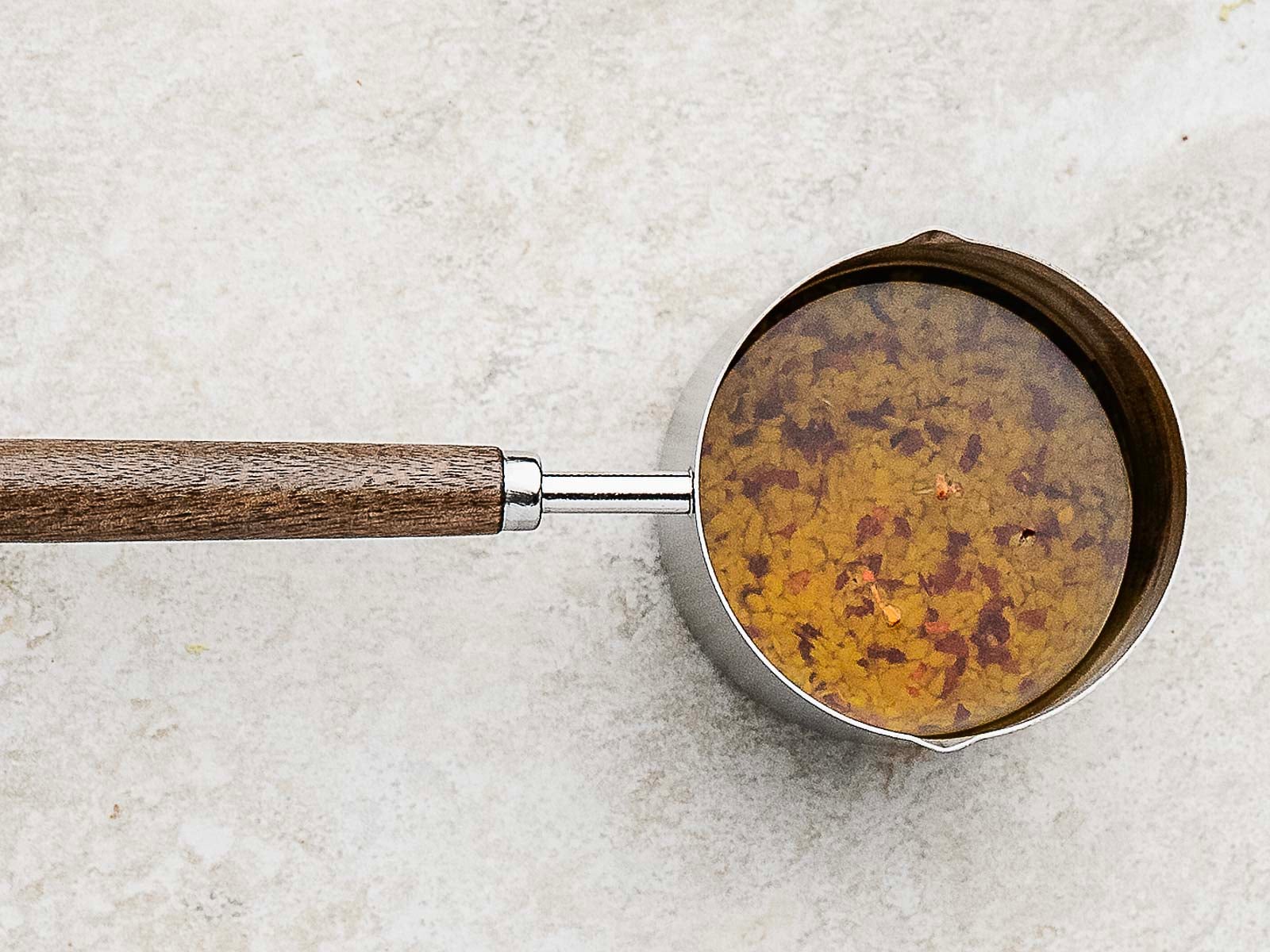 A stainless steel saucepan with a wooden handle contains a golden broth with visible herbs and spices, set on a light-colored surface.