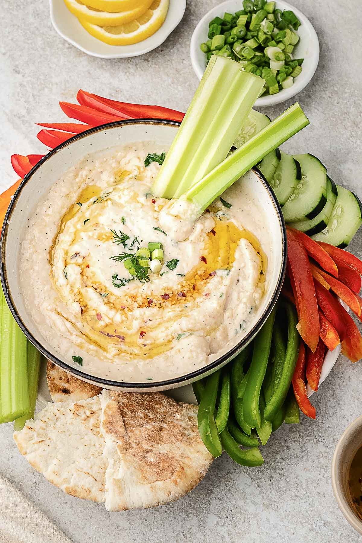 A bowl of hummus garnished with herbs and olive oil, surrounded by pita bread, celery, cucumber, and red and green bell pepper slices.