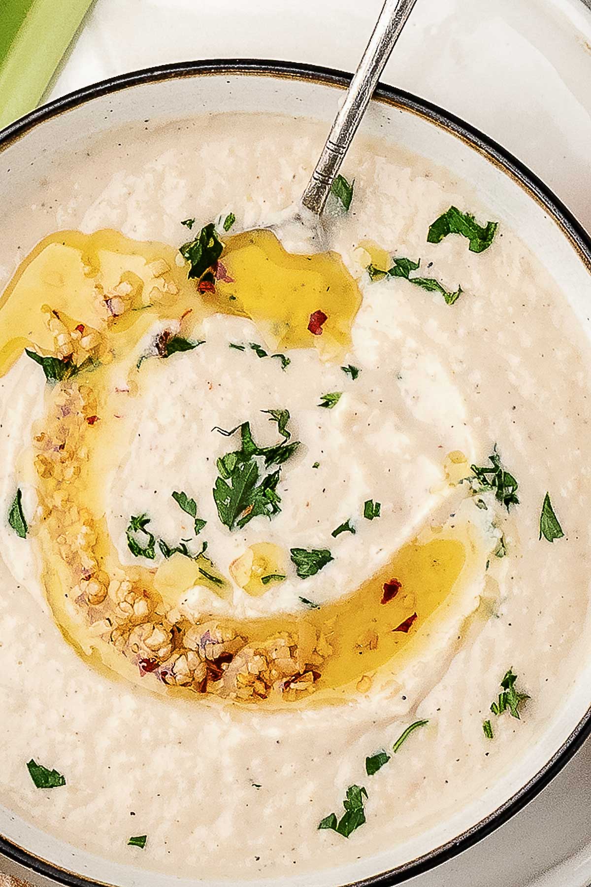 A bowl of creamy white soup garnished with chopped herbs, red chili flakes, and a swirl of olive oil, with a spoon resting inside.