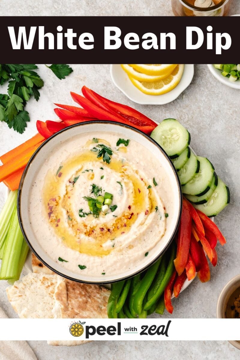 A bowl of creamy White Bean Dip topped with fresh herbs and oil, surrounded by sliced vegetables and pita bread on a light-colored surface.