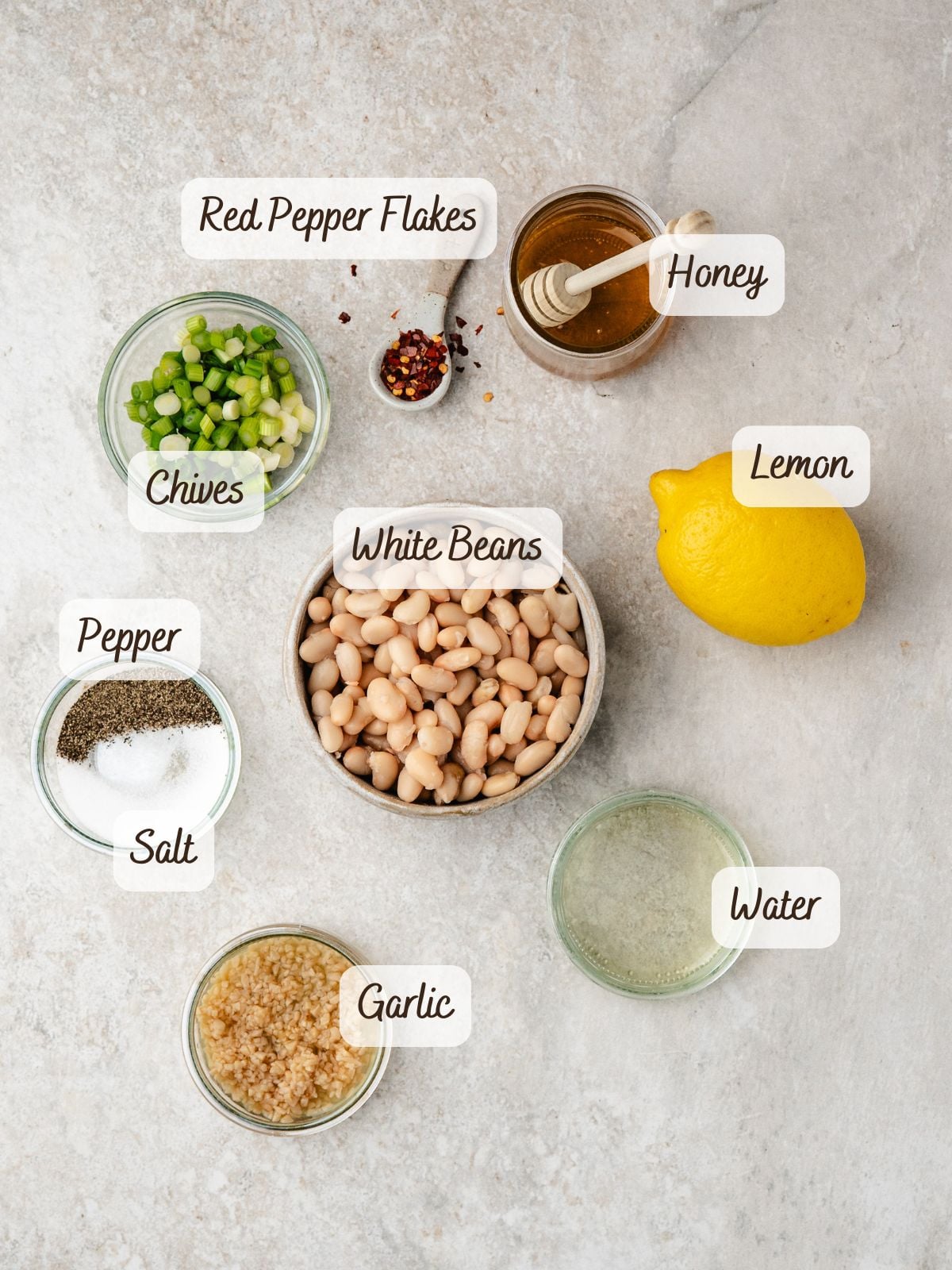 A flat lay of ingredients in bowls for White Bean Dip, including white beans, chopped chives, minced garlic, salt, pepper, red pepper flakes, honey, a lemon, and a glass of water on a light surface.