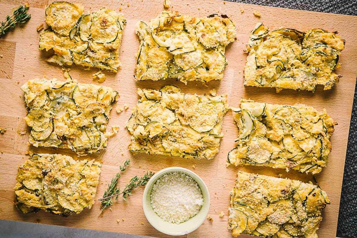 Nine rectangular pieces of zucchini casserole arranged on a wooden cutting board, with a small bowl of grated cheese and a sprig of thyme beside them.