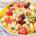 A plate of rotini pasta salad with cherry tomatoes, cucumber, red onion, Kalamata olives, feta cheese, and a pepperoncini.