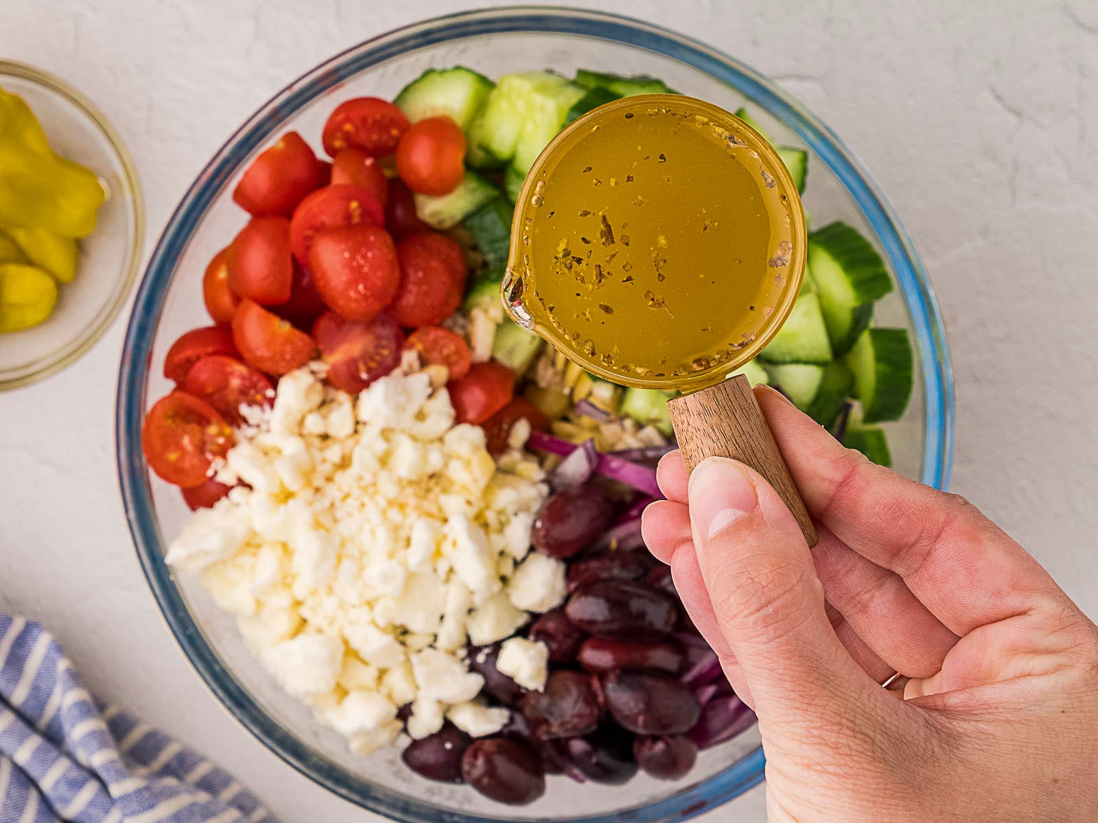 A hand holds a measuring cup of dressing above a glass bowl containing chopped tomatoes, cucumbers, onions, feta cheese, and olives.
