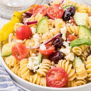 A plate of rotini pasta salad with cherry tomatoes, cucumber, red onion, Kalamata olives, feta cheese, and a pepperoncini.