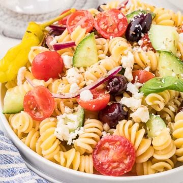A plate of rotini pasta salad with cherry tomatoes, cucumber, red onion, Kalamata olives, feta cheese, and a pepperoncini.