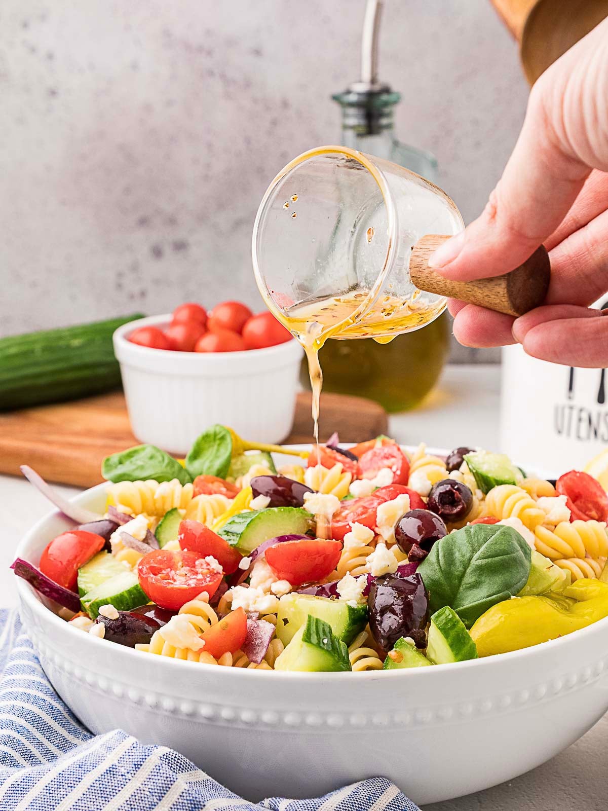 A hand pours dressing onto a bowl of pasta salad with cucumbers, tomatoes, olives, and feta cheese. A jar of olive oil and a bowl of tomatoes are in the background.