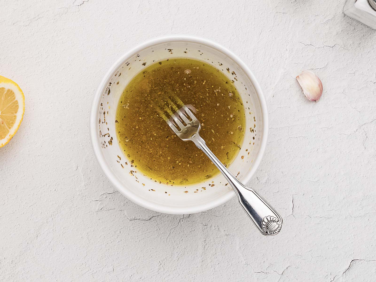 A white bowl filled with yellow vinaigrette dressing and a fork, placed on a light surface with a lemon wedge and a garlic clove nearby.