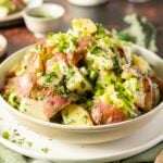 A bowl of potato salad made with red-skinned potatoes, chopped green herbs, and celery, served on a white plate.