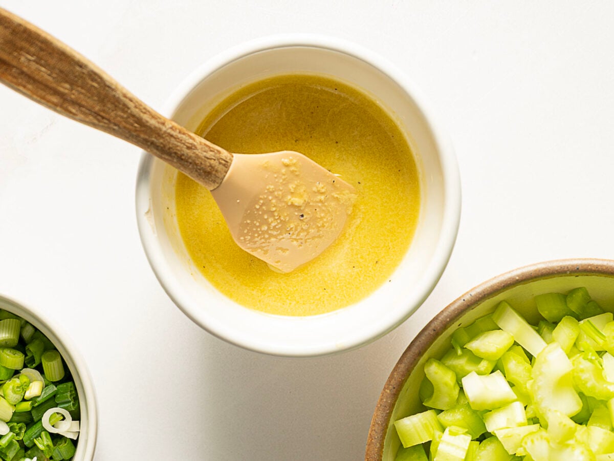 A bowl of yellow dressing with a spatula, next to bowls of chopped green onions and chopped celery on a white surface.