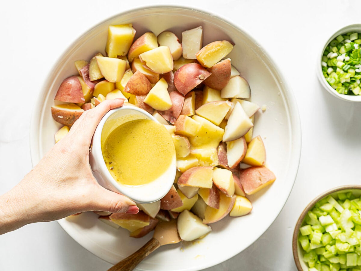 A hand pours yellow dressing over a bowl of chopped red potatoes, with bowls of chopped celery and green onions nearby.