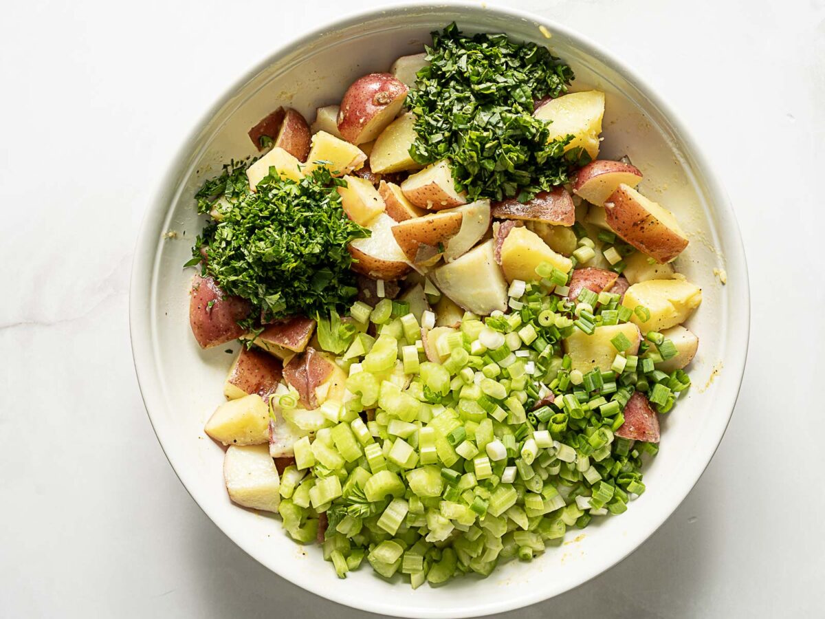 A white bowl contains chopped red potatoes, celery, green onions, and fresh herbs on a white surface.