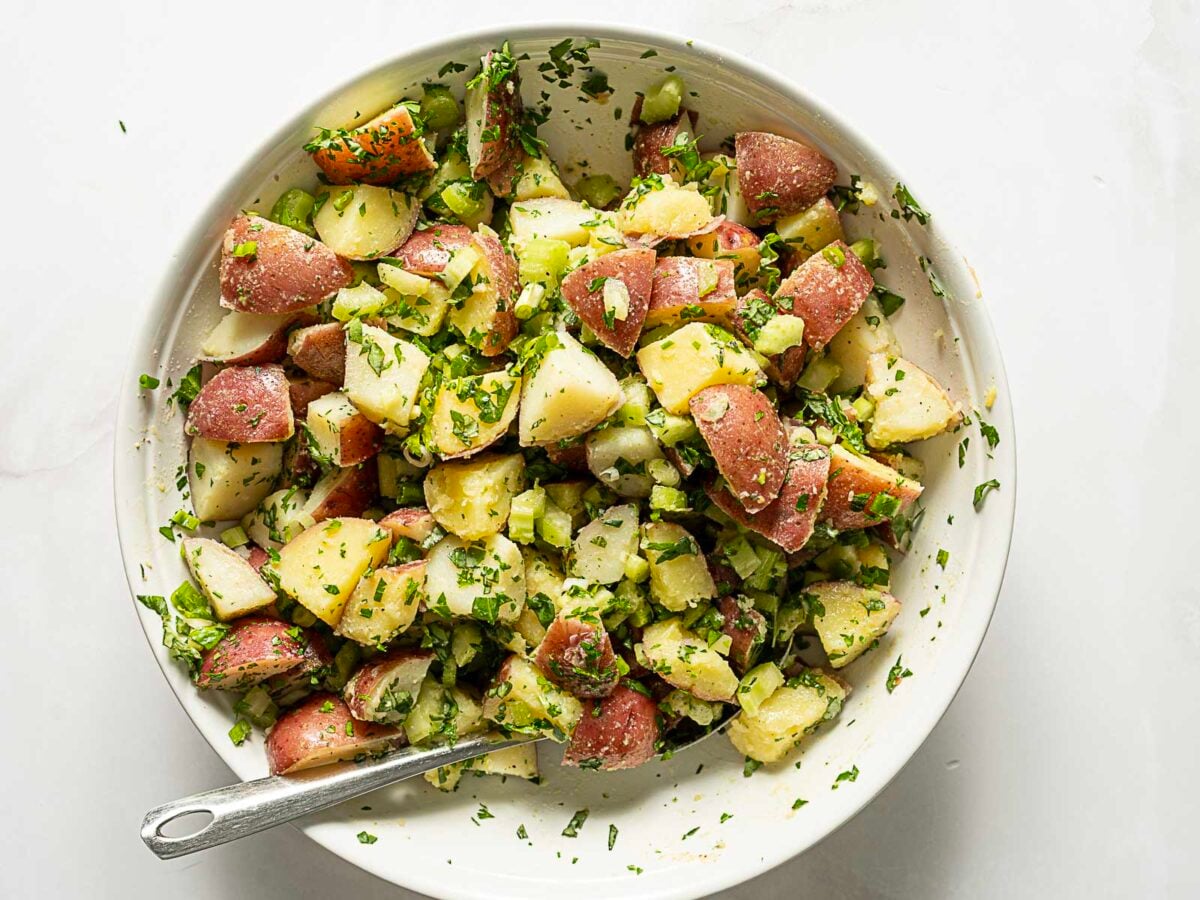 A bowl of potato salad made with red potatoes, chopped celery, and fresh herbs, with a metal spoon resting inside.