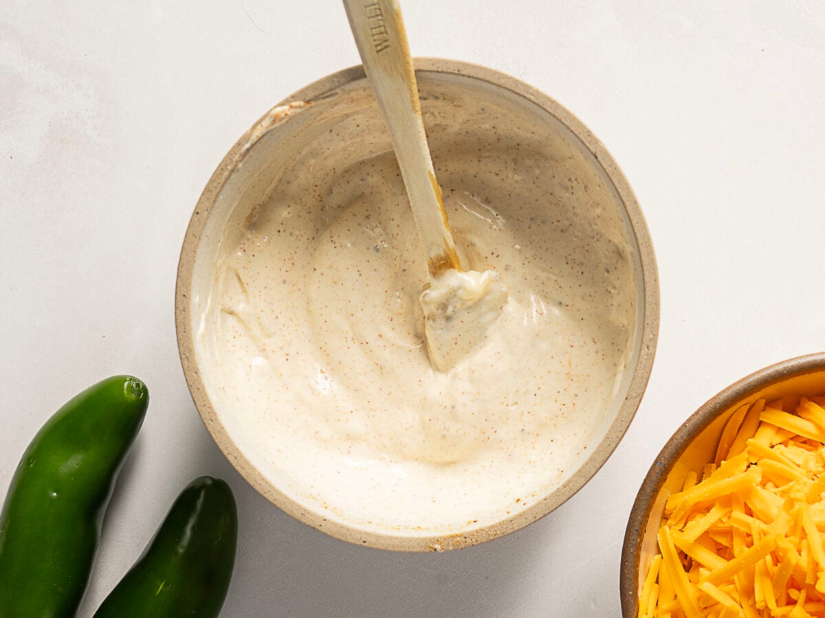 A bowl of creamy white sauce is being stirred with a wooden spoon, with jalape&ntilde;os and shredded cheddar cheese nearby on a white surface.