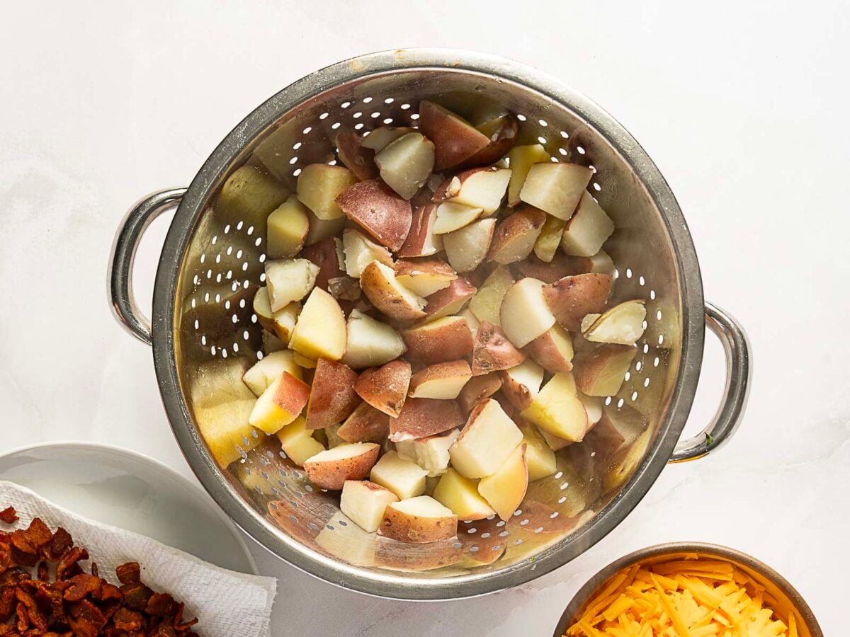 A metal colander filled with steamed, chopped red potatoes sits on a white surface beside bowls of bacon and shredded cheese.