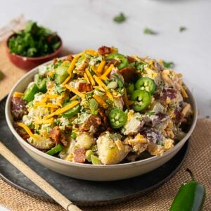 A bowl of loaded potato salad topped with shredded cheddar cheese, sliced jalape&ntilde;os, bacon, and chopped herbs on a table with a small bowl of greens in the background.