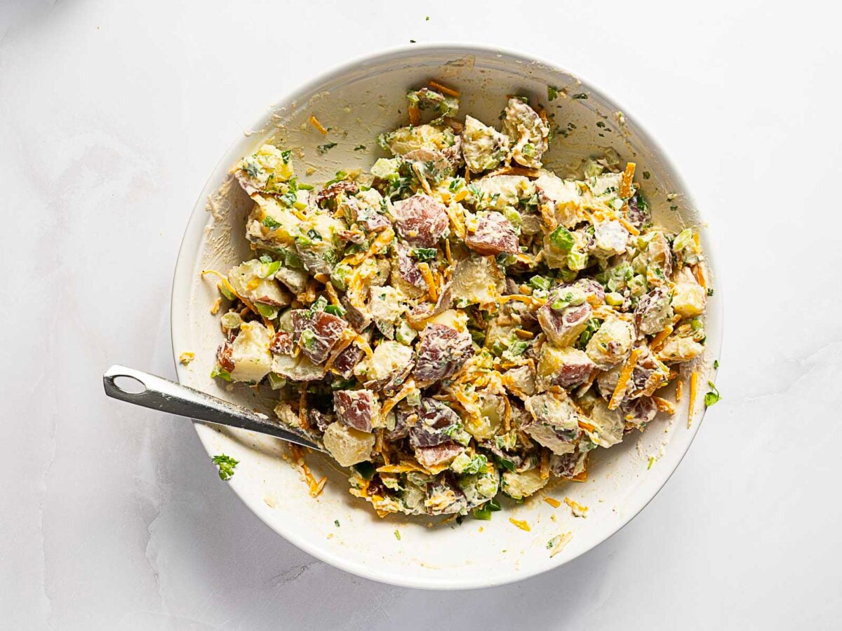 A white bowl filled with mixed potato salad containing red potatoes, herbs, and a creamy dressing, with a metal spoon resting inside.