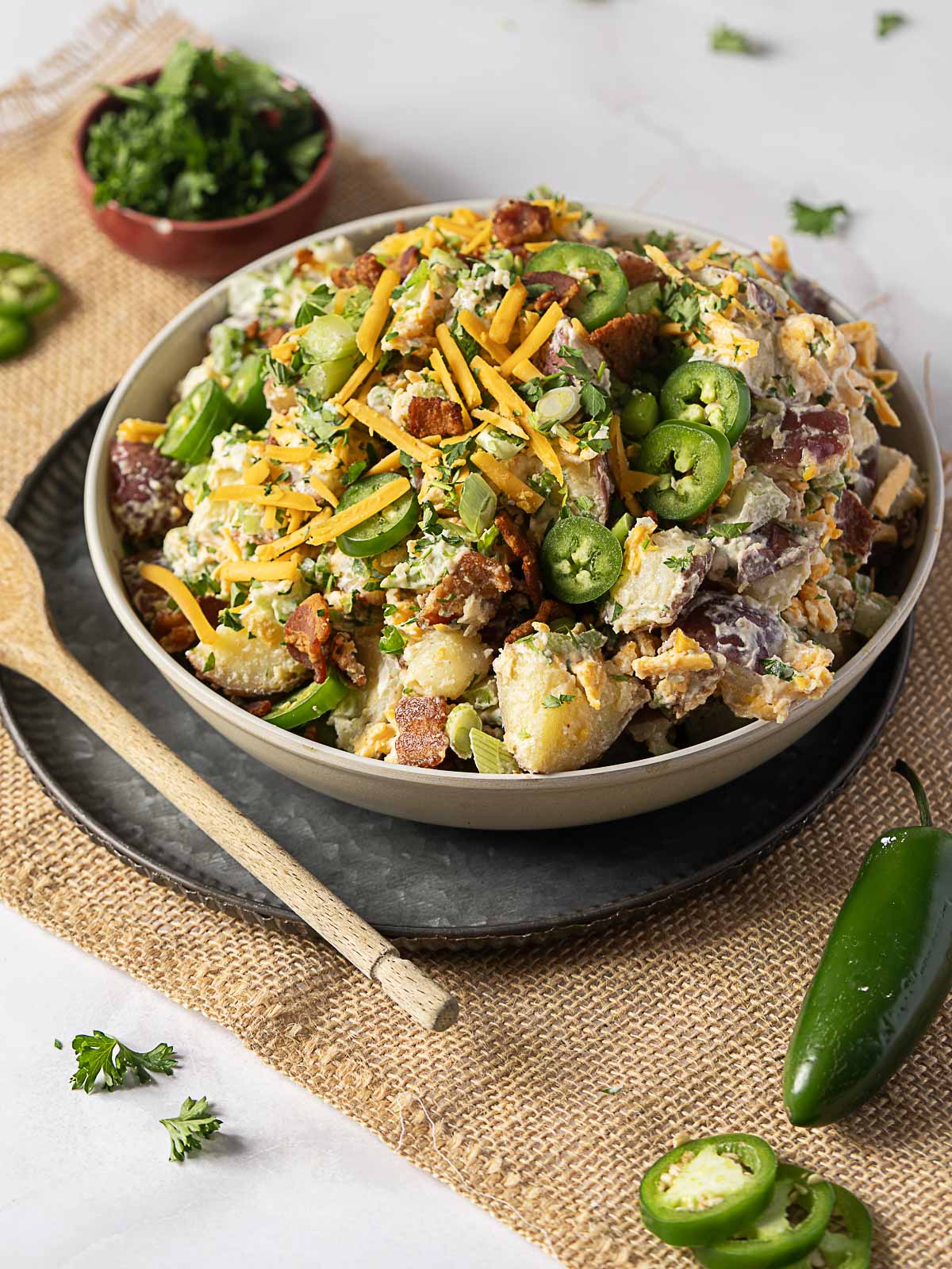 A bowl of loaded potato salad topped with shredded cheddar cheese, sliced jalape&ntilde;os, bacon bits, and chopped herbs, with a wooden spoon and fresh jalape&ntilde;o on the side.
