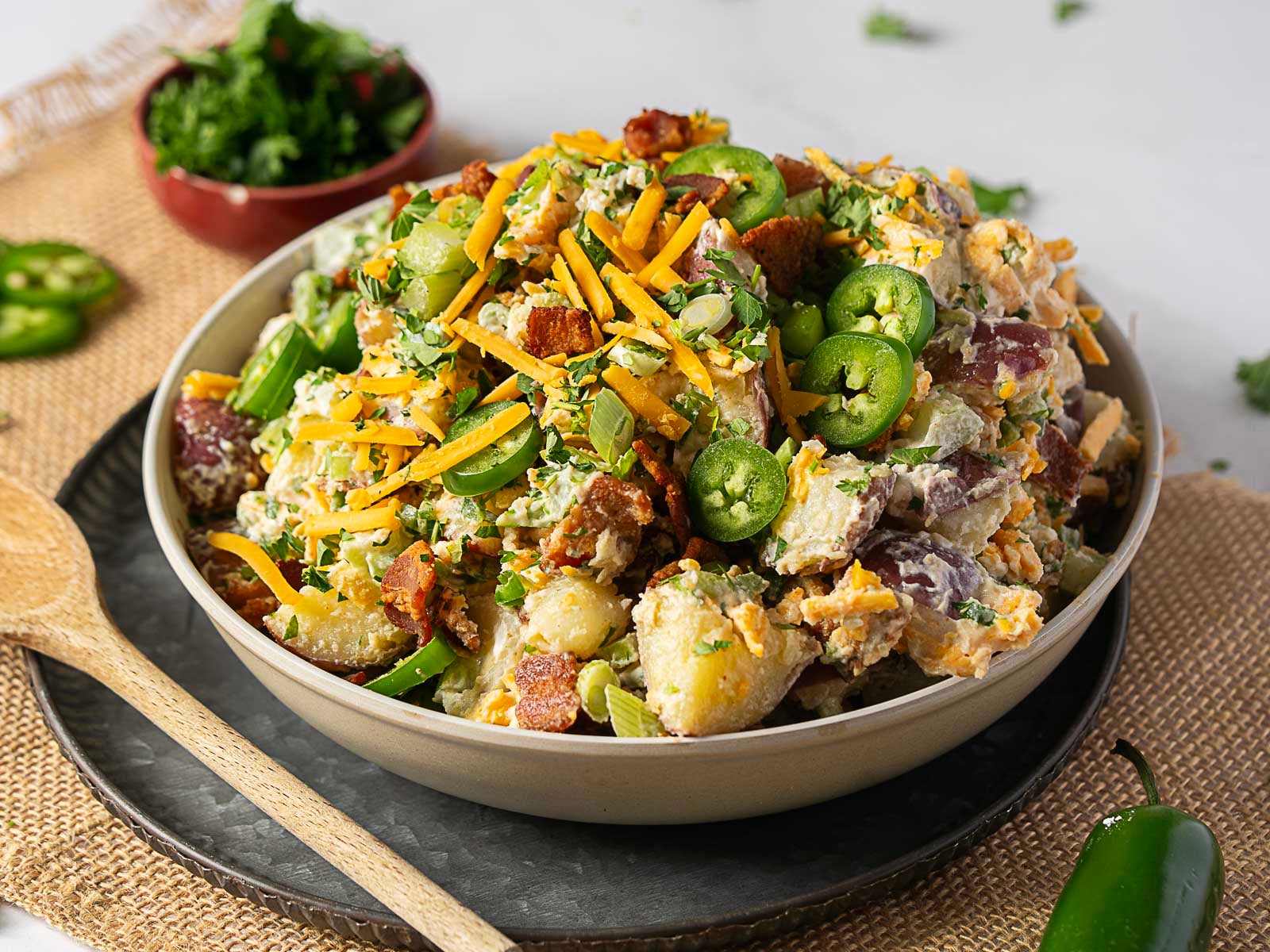 A bowl of potato salad topped with shredded cheddar cheese, bacon bits, sliced jalapeños, and chopped herbs sits on a plate beside a wooden spoon.