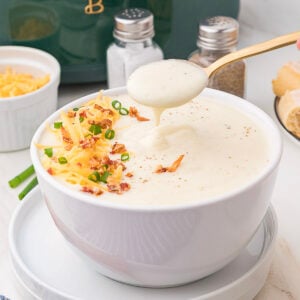 A bowl of creamy soup garnished with shredded cheese, bacon bits, and chopped green onions; a spoon lifts some soup out of the bowl.