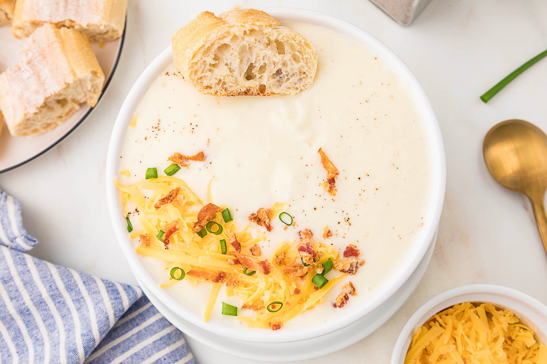 A bowl of cheesy cauliflower soup topped with shredded cheese, bacon bits, and chopped chives, with a piece of bread on the rim; bread slices and a gold spoon are nearby.