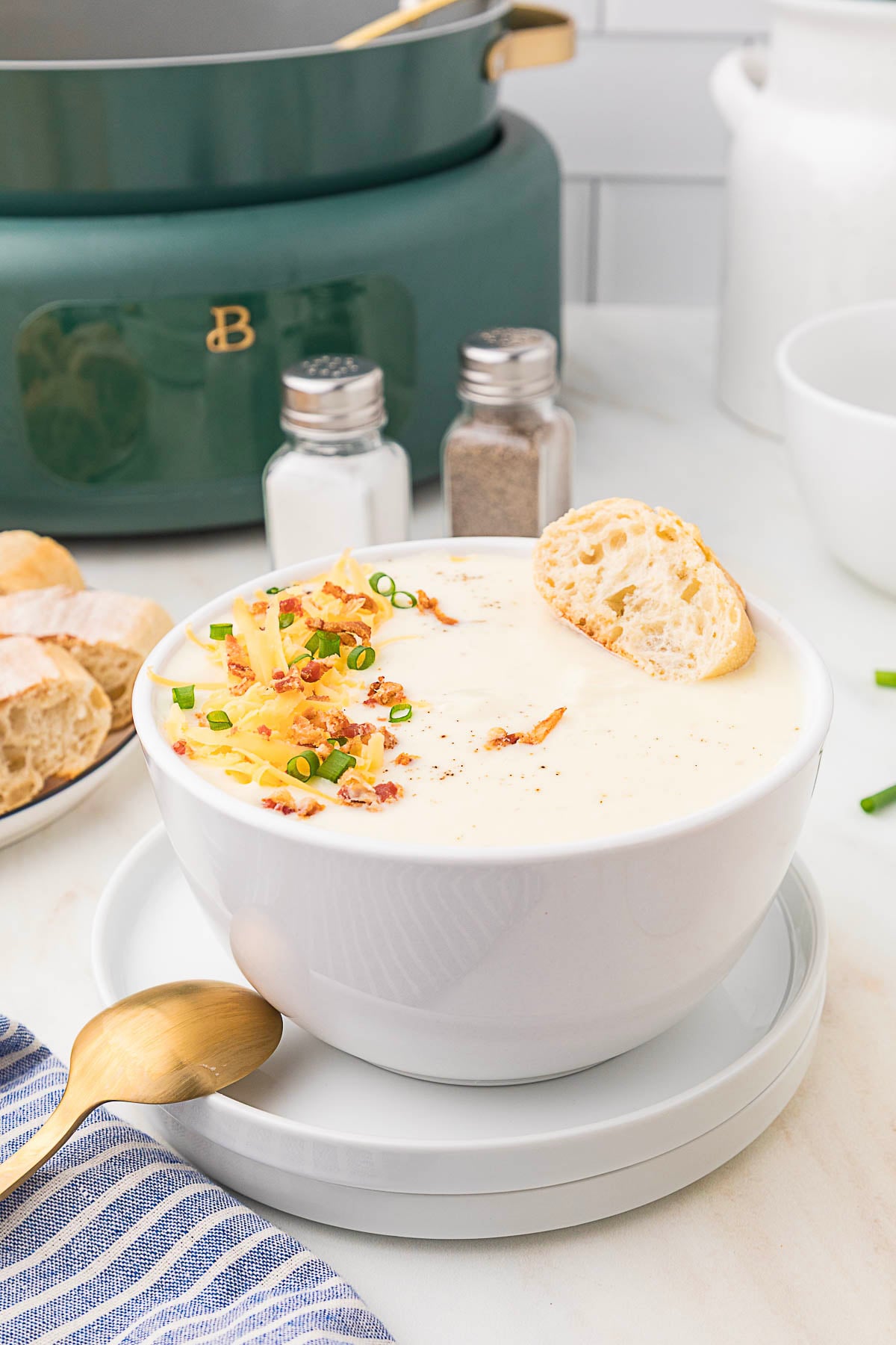 A bowl of cheesy cauliflower soup, garnished with shredded cheese, bacon bits, and chopped green onions, with a slice of bread on the rim, sits on a white plate beside a gold spoon. Perfect for slow cooker meal prep.