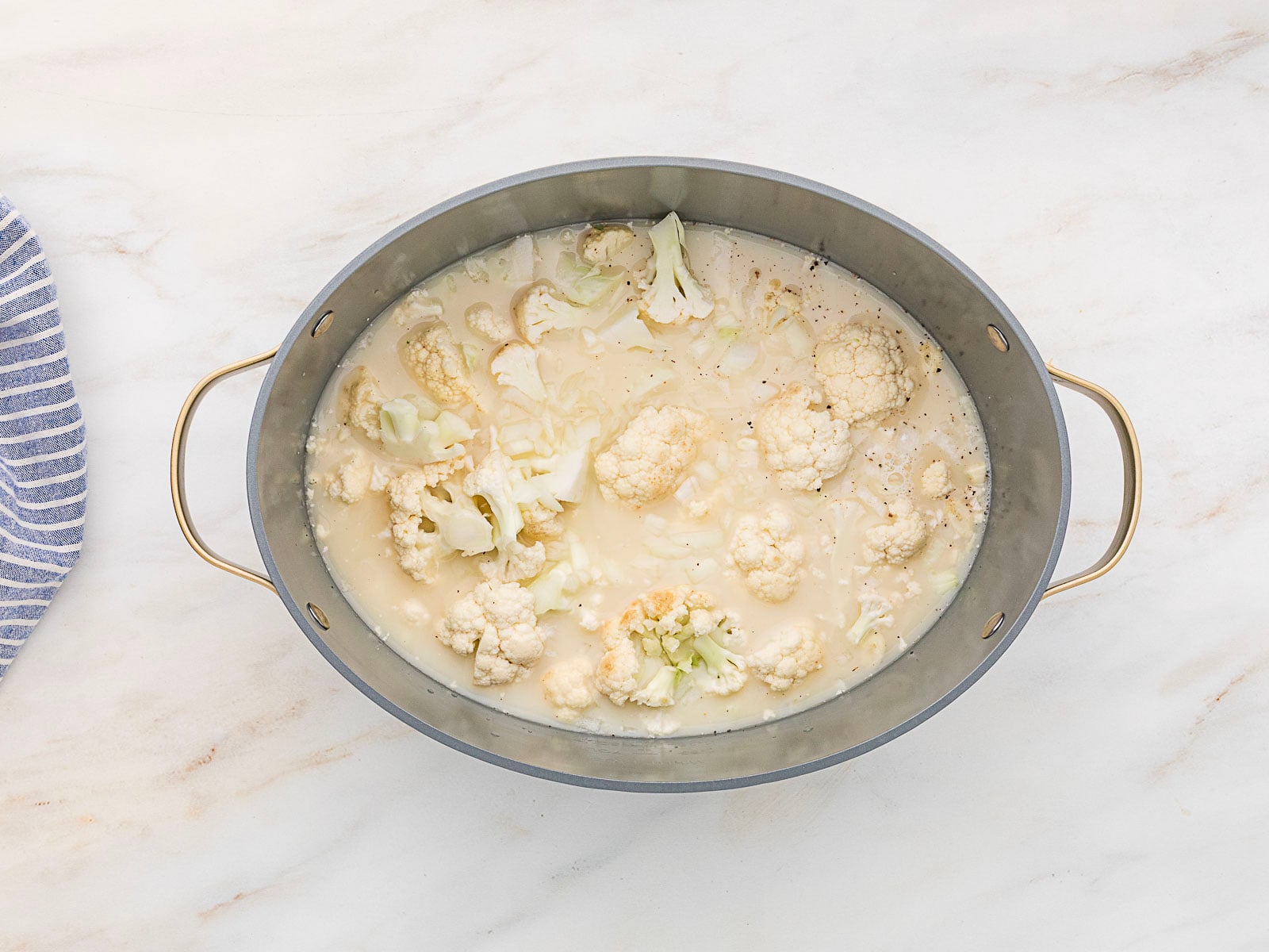 Oval pot with brass handles containing cauliflower florets in a creamy, light-colored liquid. The pot is on a marble countertop next to a striped cloth.