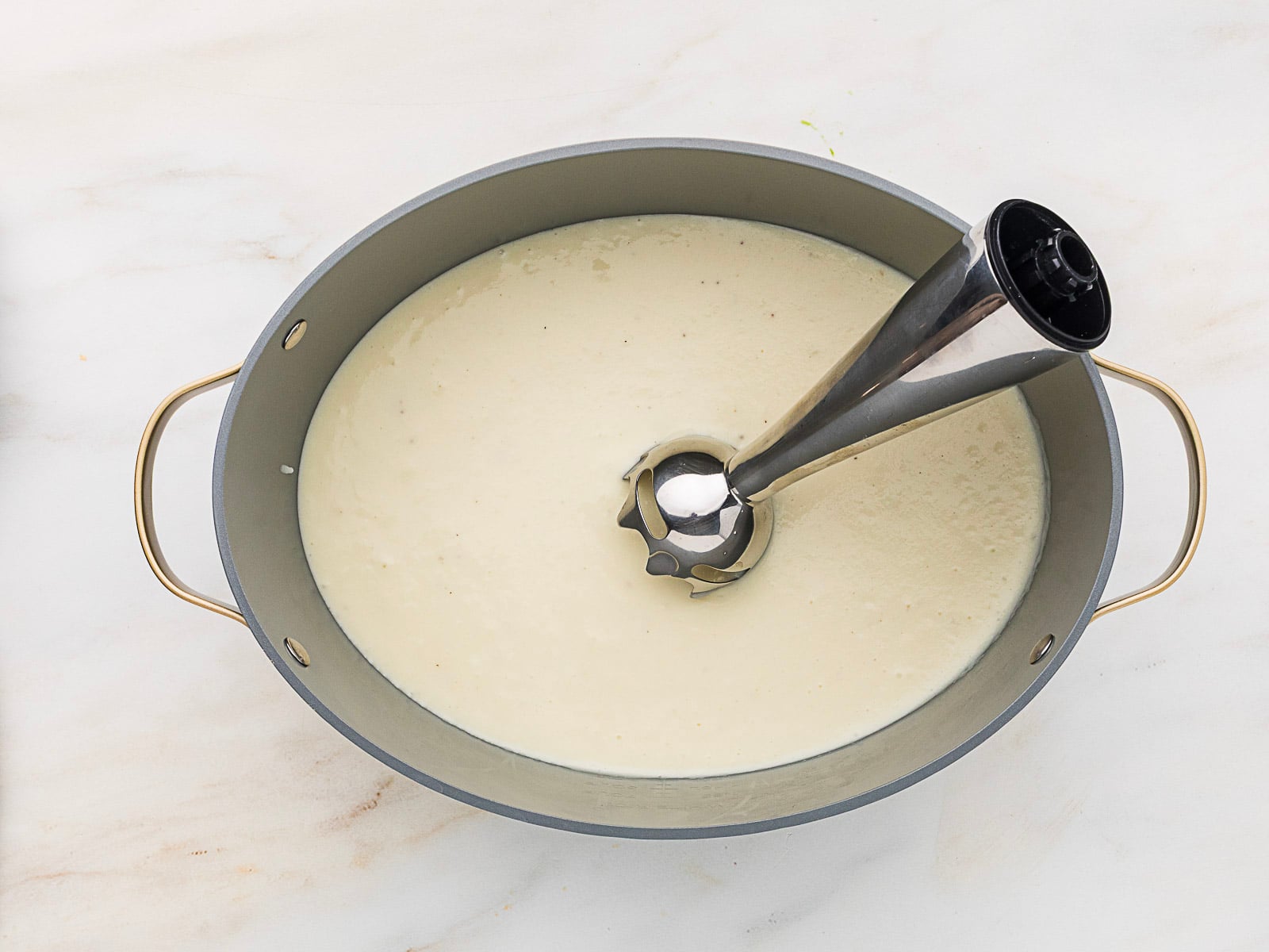 Oval pan filled with creamy white soup or sauce, with an immersion blender resting inside on a white marble surface.