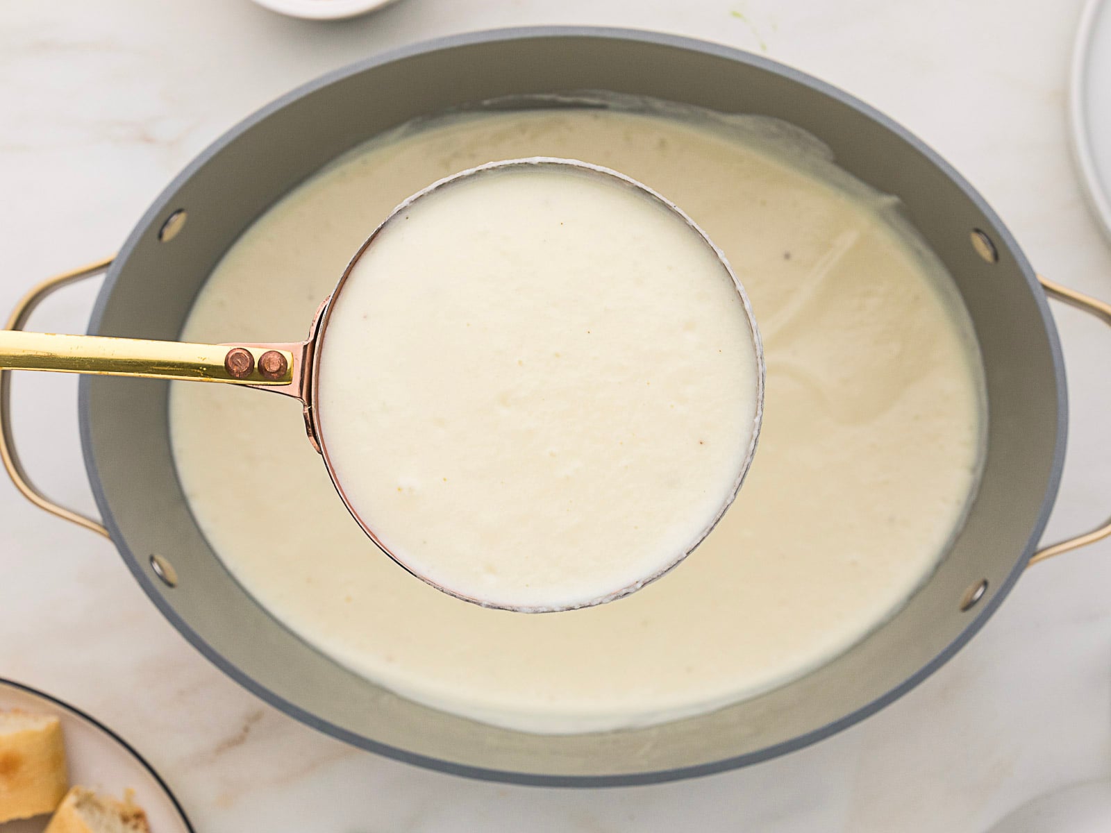 A ladle holding a creamy white sauce above a gray pot filled with the same sauce, on a light countertop.