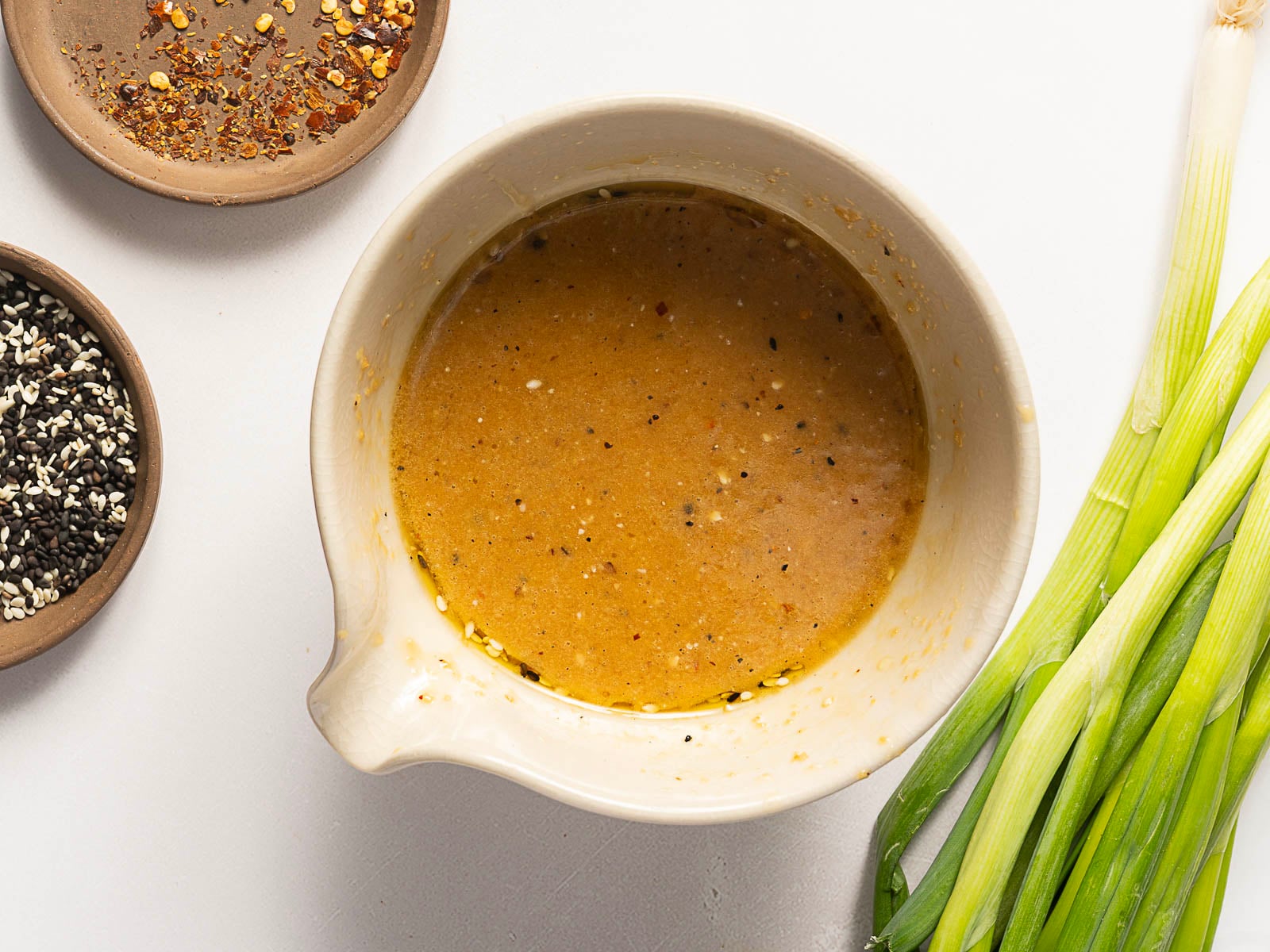 A bowl of light brown sauce sits on a white surface next to green onions, a small dish of sesame seeds, and a small dish of red pepper flakes.