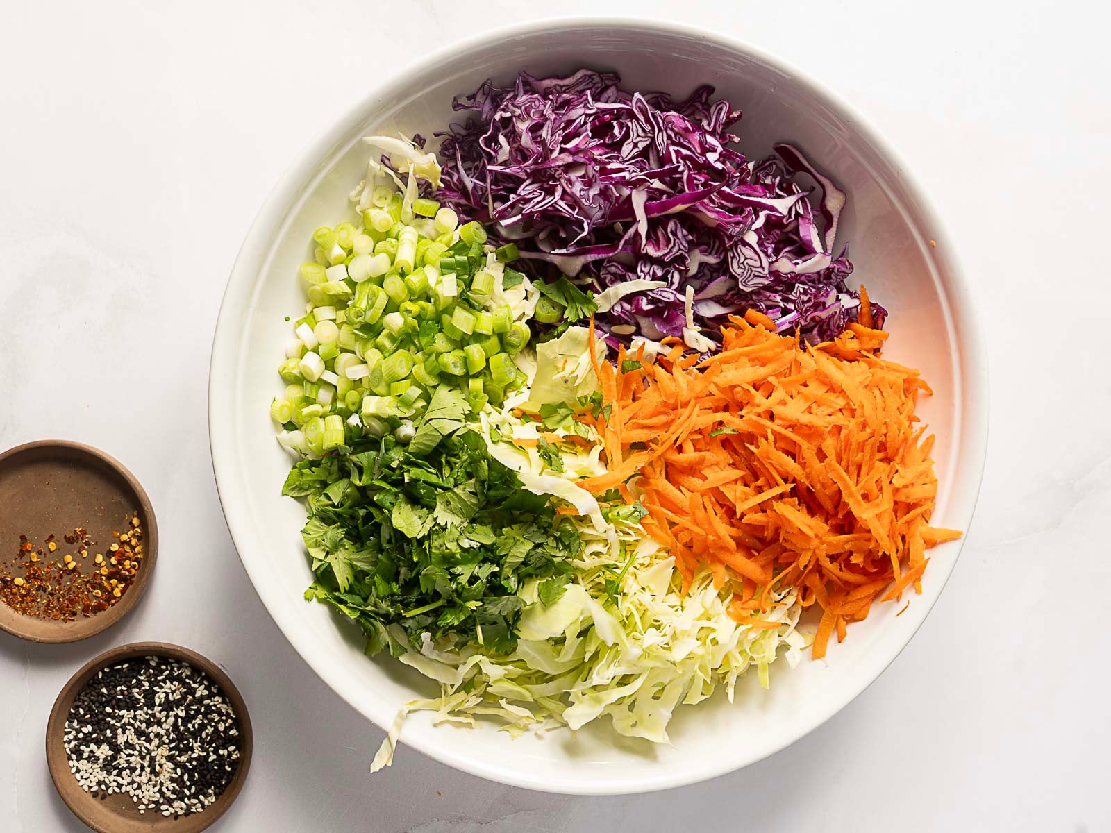 A white bowl containing shredded purple cabbage, shredded carrots, chopped green onions, chopped cilantro, and shredded green cabbage. Two small bowls with spices are beside it.