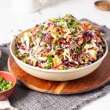 A bowl of mixed coleslaw with shredded cabbage, carrots, and herbs sits on a wooden board with a small dish of chopped green onions nearby.