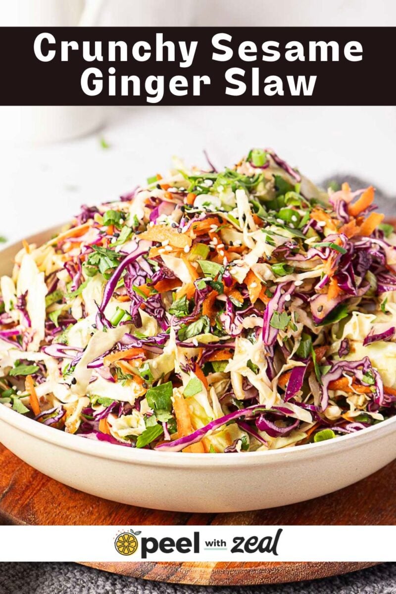 A bowl of crunchy sesame ginger slaw with shredded cabbage, carrots, and herbs, sitting on a wooden surface.
