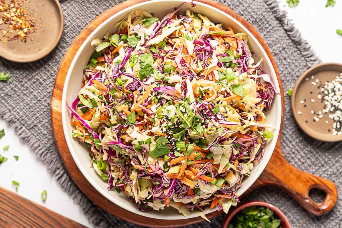 A bowl of colorful cabbage slaw with shredded purple and green cabbage, carrots, cilantro, and sesame seeds, placed on a wooden tray with small dishes of seasoning nearby.